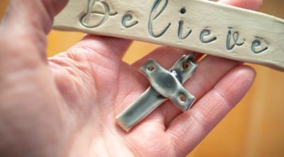 A hand holding a pottery cross and plaque with the word "Believe."