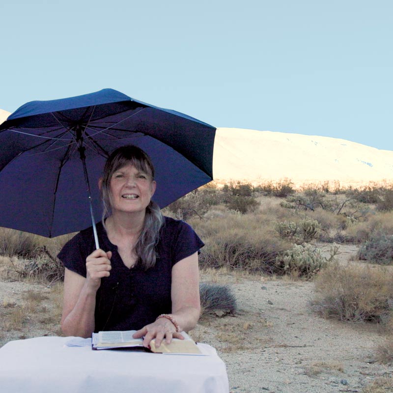 Josephine Mary Schmidt sitting at a table with a white tablecloth holding an umbrella in the desert in Grapevine Canyon