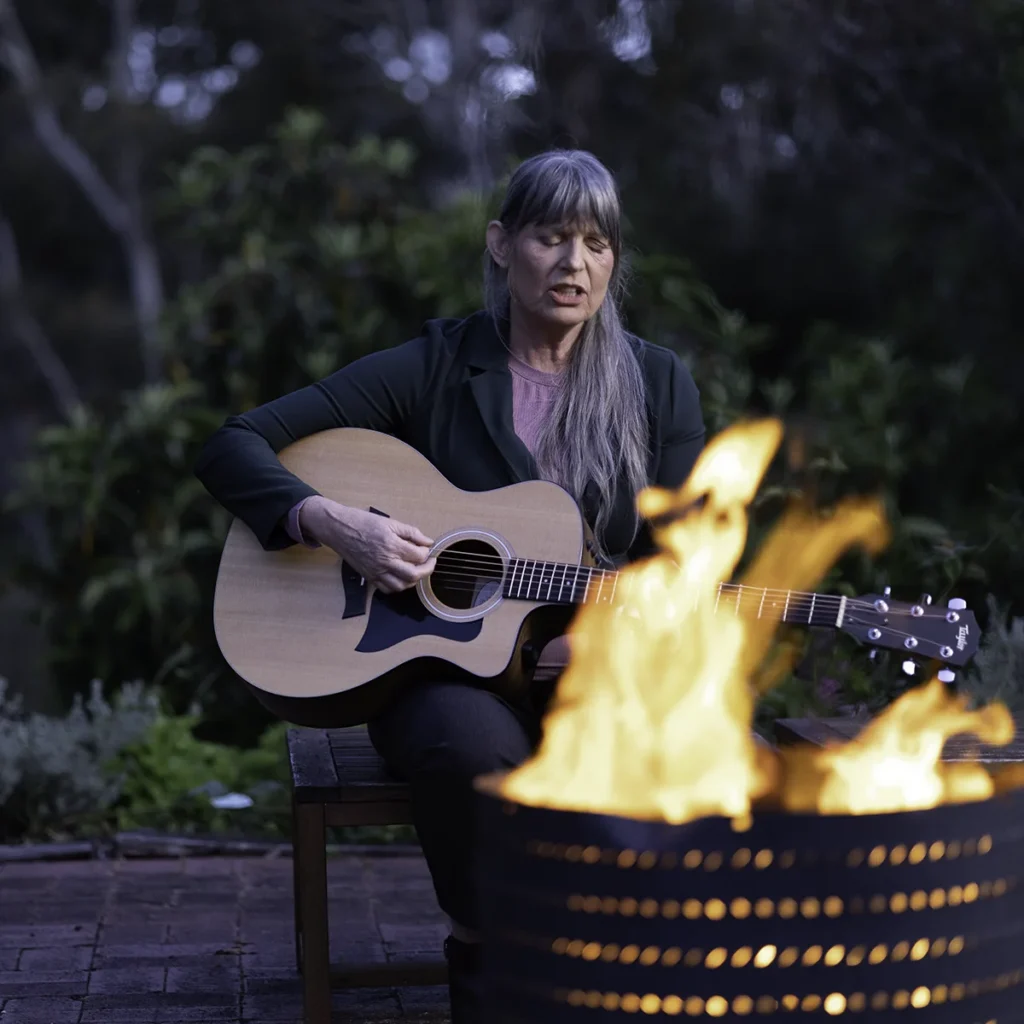 Josephine Mary Schmidt is singing sincerely, playing her acoustic guitar outside next to an open pit fire with flames