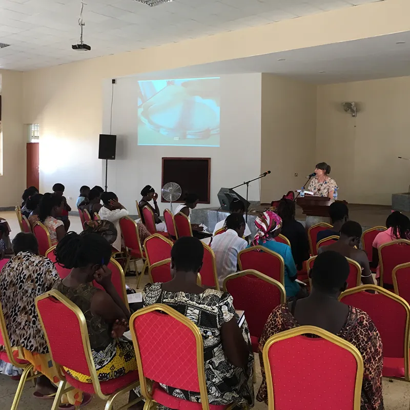 Josephine Mary Schmidt speaking into a microphone with her bible teaching women in Soroti, Uganda, with a pottery plate image projected on the wall